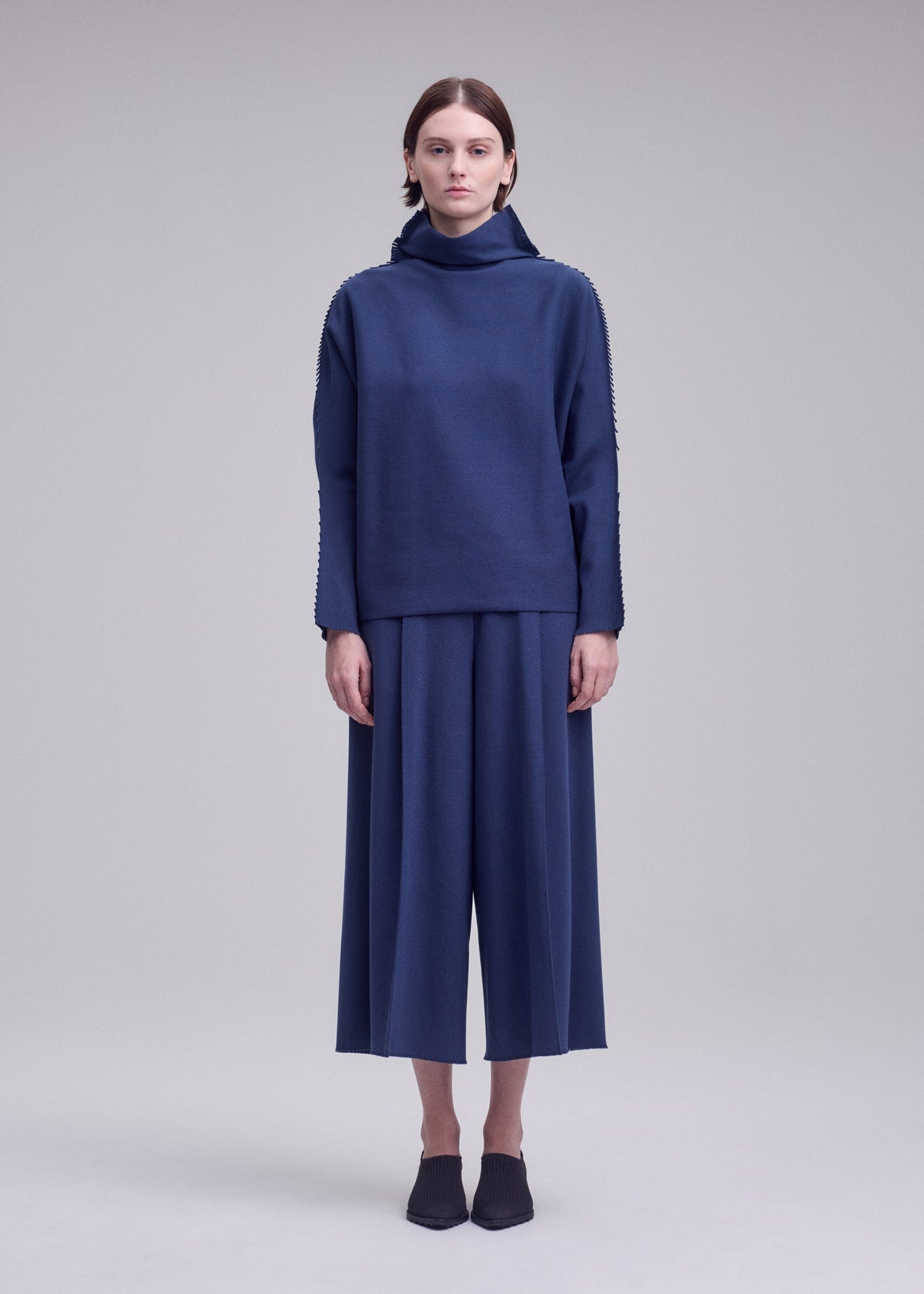 A person stands facing forward, wearing a loose-fitting, long-sleeved polyurethane-blend blue top and wide-legged blue culottes. Their hair is pulled back, and they wear black pointed shoes against a plain light background. view 3 of 5