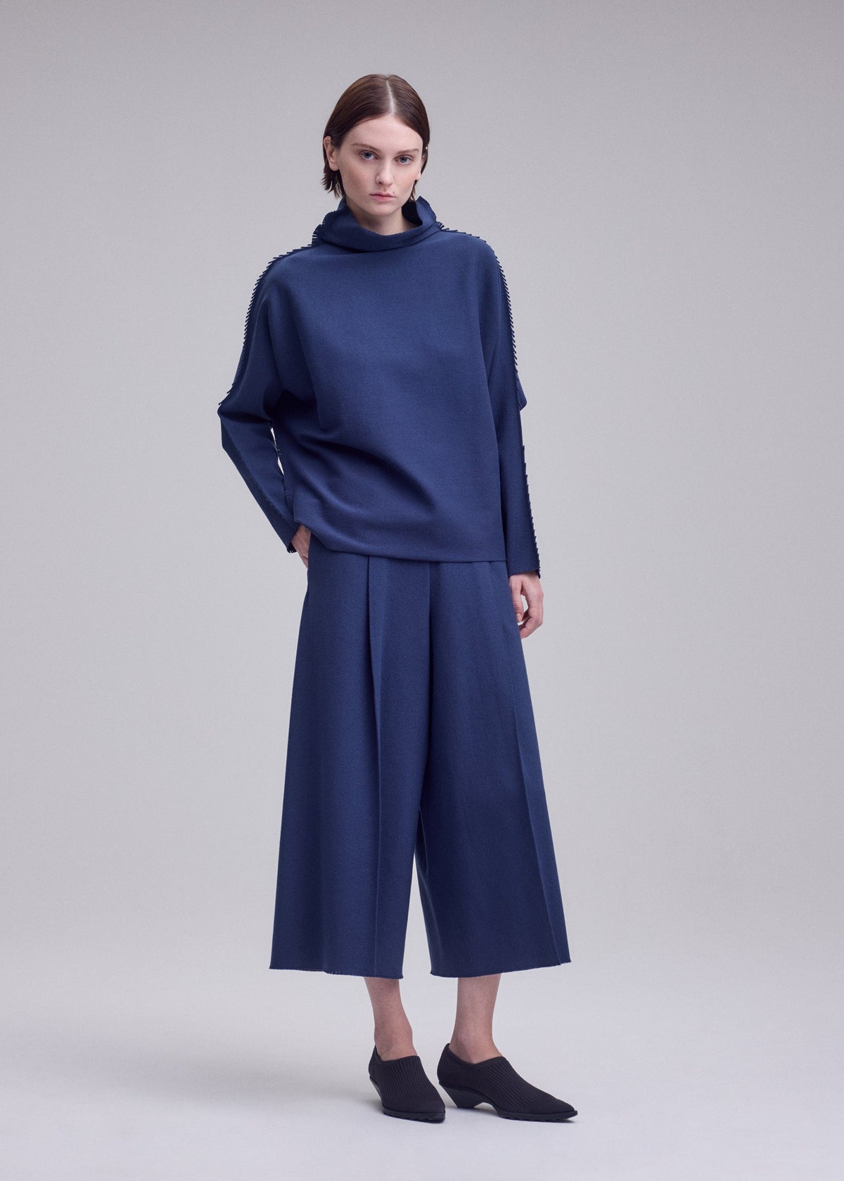 A person stands against a plain light background wearing the CAMPAGNE ensemble by ISSEY MIYAKE: a loose-fitting, long-sleeved navy cowl-neck top and matching wide-leg culottes crafted from a soft polyurethane-blend fabric, paired with black slip-on shoes. view 4 of 5