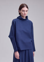 A person with short brown hair wears the CAMPAGNE loose-fitting, long-sleeved polyurethane-blend top and matching skirt by ISSEY MIYAKE, standing against a plain light gray background.
