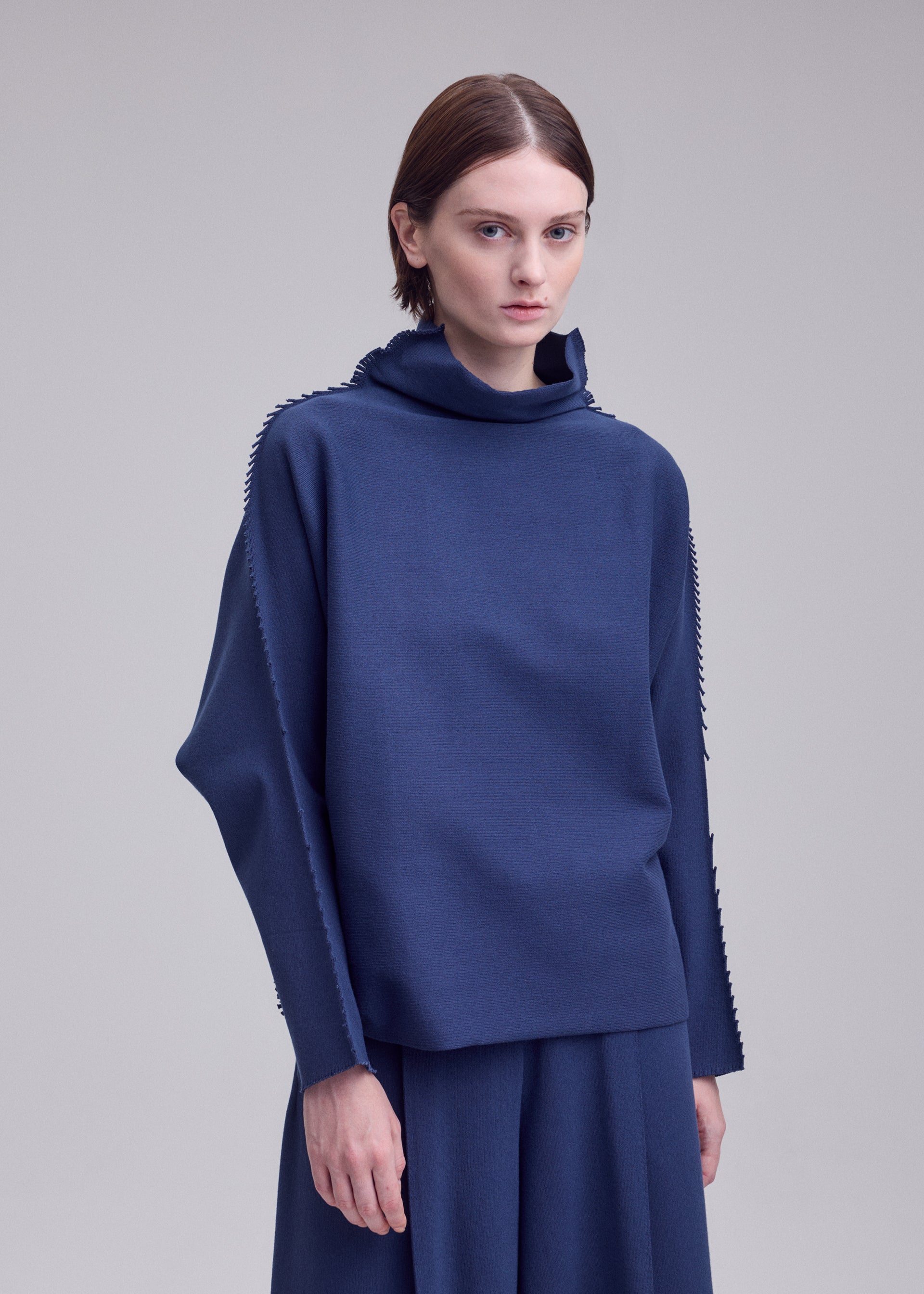 Zoomed view A person with short brown hair wears the CAMPAGNE loose-fitting, long-sleeved polyurethane-blend top and matching skirt by ISSEY MIYAKE, standing against a plain light gray background.