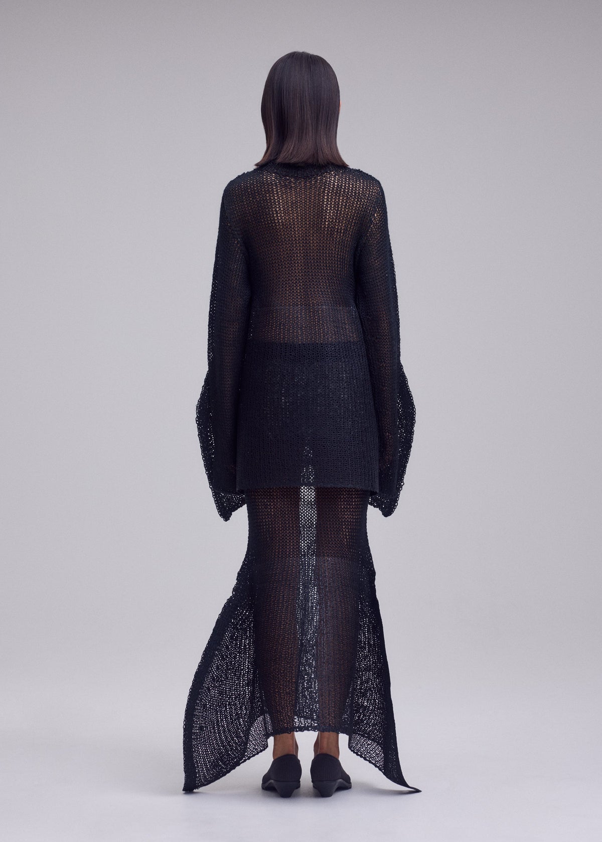A person faces away, wearing the ISSEY MIYAKE PANINI PULLED—a sheer black dress with wide sleeves—layered over a long-sleeved wool-alpaca knit top and black shoes against a light, plain background. view 6 of 7