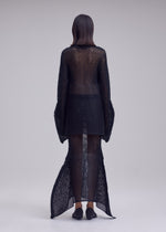 A person faces away, wearing the ISSEY MIYAKE PANINI PULLED—a sheer black dress with wide sleeves—layered over a long-sleeved wool-alpaca knit top and black shoes against a light, plain background.
