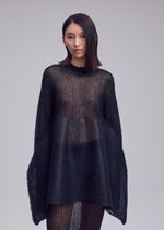 A woman with straight, shoulder-length hair models the PANINI PULLED dress by ISSEY MIYAKE—a loose black knitwear piece styled over a skirt, set against a plain light background.