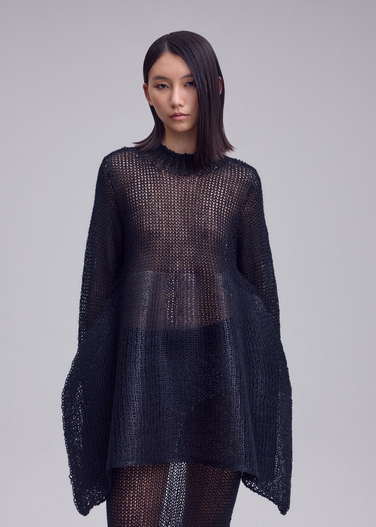 Zoomed view A woman with straight, shoulder-length hair models the PANINI PULLED dress by ISSEY MIYAKE—a loose black knitwear piece styled over a skirt, set against a plain light background.