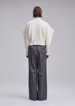 A person faces away, wearing the ISSEY MIYAKE O KNIT with asymmetrical sleeves and loose gray trousers in 100% wool yarn, set against a plain light background.