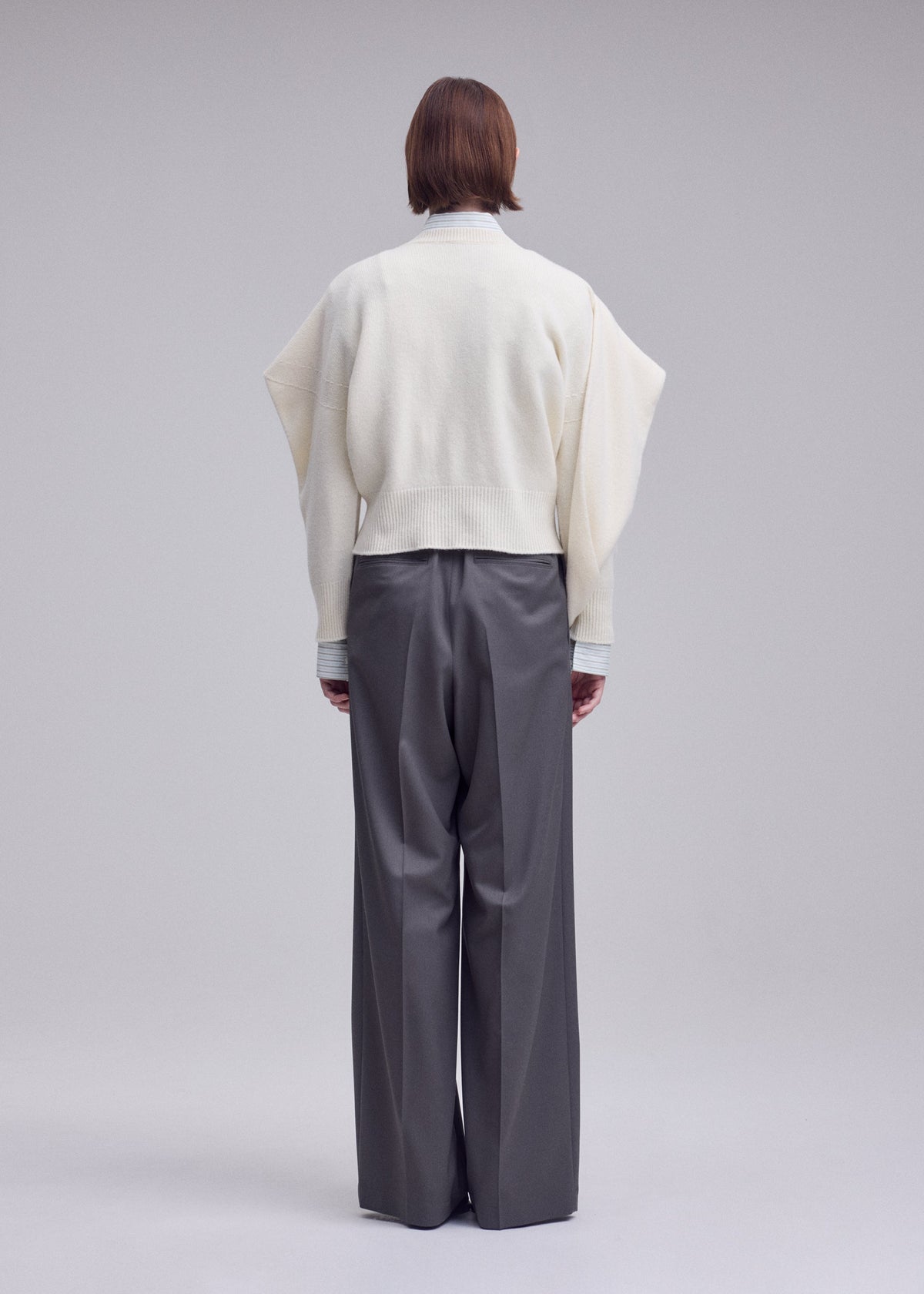 Zoomed view A person faces away, wearing the ISSEY MIYAKE O KNIT with asymmetrical sleeves and loose gray trousers in 100% wool yarn, set against a plain light background.
