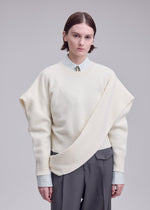 A person with short brown hair wears the ISSEY MIYAKE O KNIT cream asymmetrical sweater over a striped shirt and tie, paired with gray pants, standing against a plain light background.