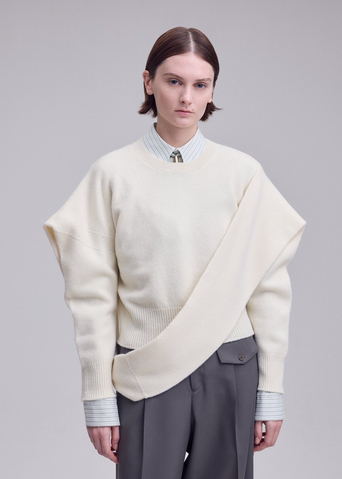 Zoomed view A person with short brown hair wears the ISSEY MIYAKE O KNIT cream asymmetrical sweater over a striped shirt and tie, paired with gray pants, standing against a plain light background.