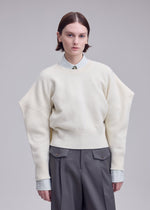 A person with short brown hair wears an ISSEY MIYAKE O KNIT over a collared shirt and gray pants, standing against a plain gray background with a neutral expression.