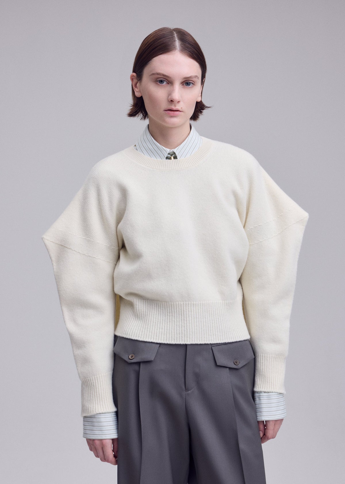 Zoomed view A person with short brown hair wears an ISSEY MIYAKE O KNIT over a collared shirt and gray pants, standing against a plain gray background with a neutral expression.