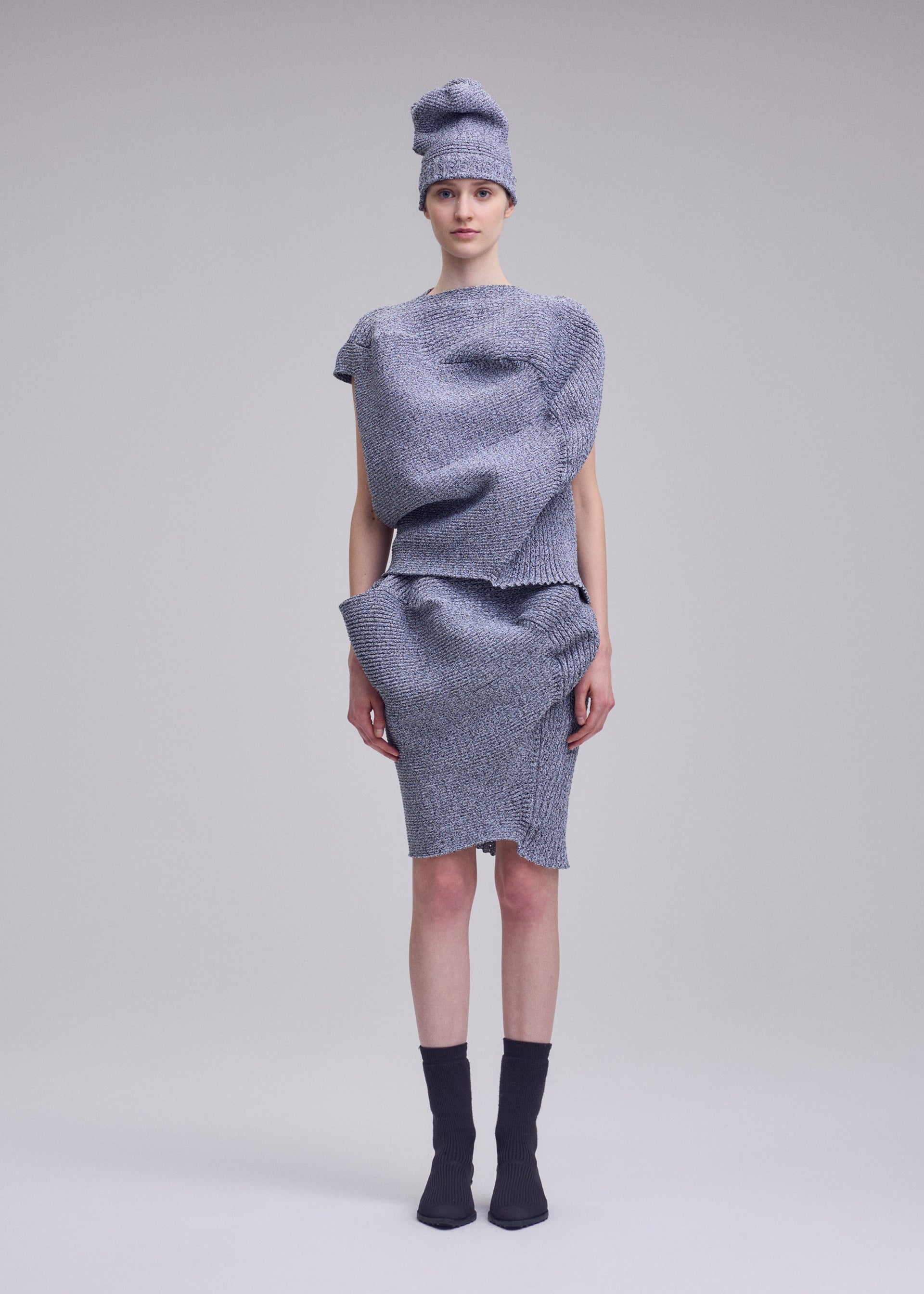 Zoomed view A person stands against a plain background wearing ISSEY MIYAKE's FOUND STONE: an asymmetrical gray organic knitwear outfit with sculptural, layered textures that evoke natural forms, completed with a matching hat, black socks, and pointed shoes.
