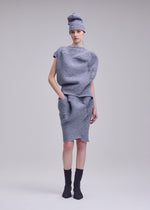 A model poses against a plain background wearing the FOUND STONE mesh-pattern top and skirt set by ISSEY MIYAKE, paired with a matching hat and black ankle socks, her hand on her hip and expression neutral.