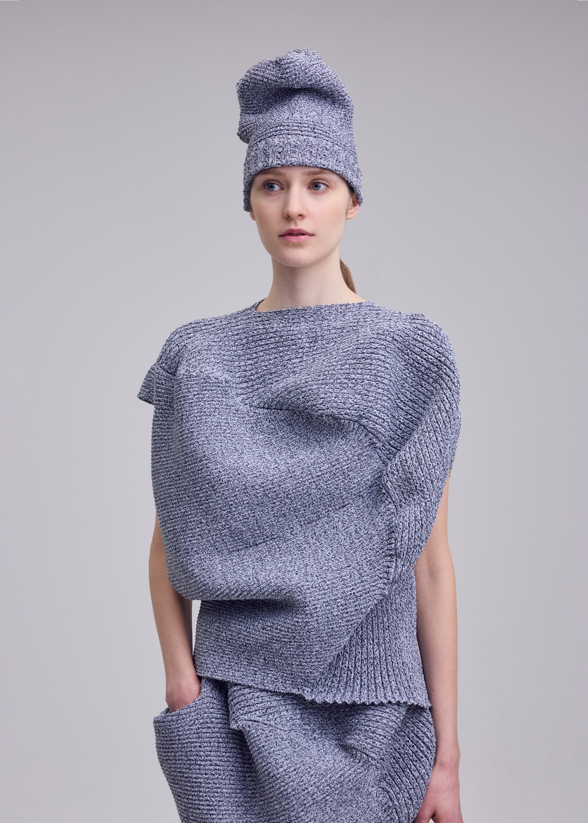 Zoomed view A person models the ISSEY MIYAKE FOUND STONE textured, stone-inspired knit top with sculptural shapes and a matching hat, posing against a plain light gray background.