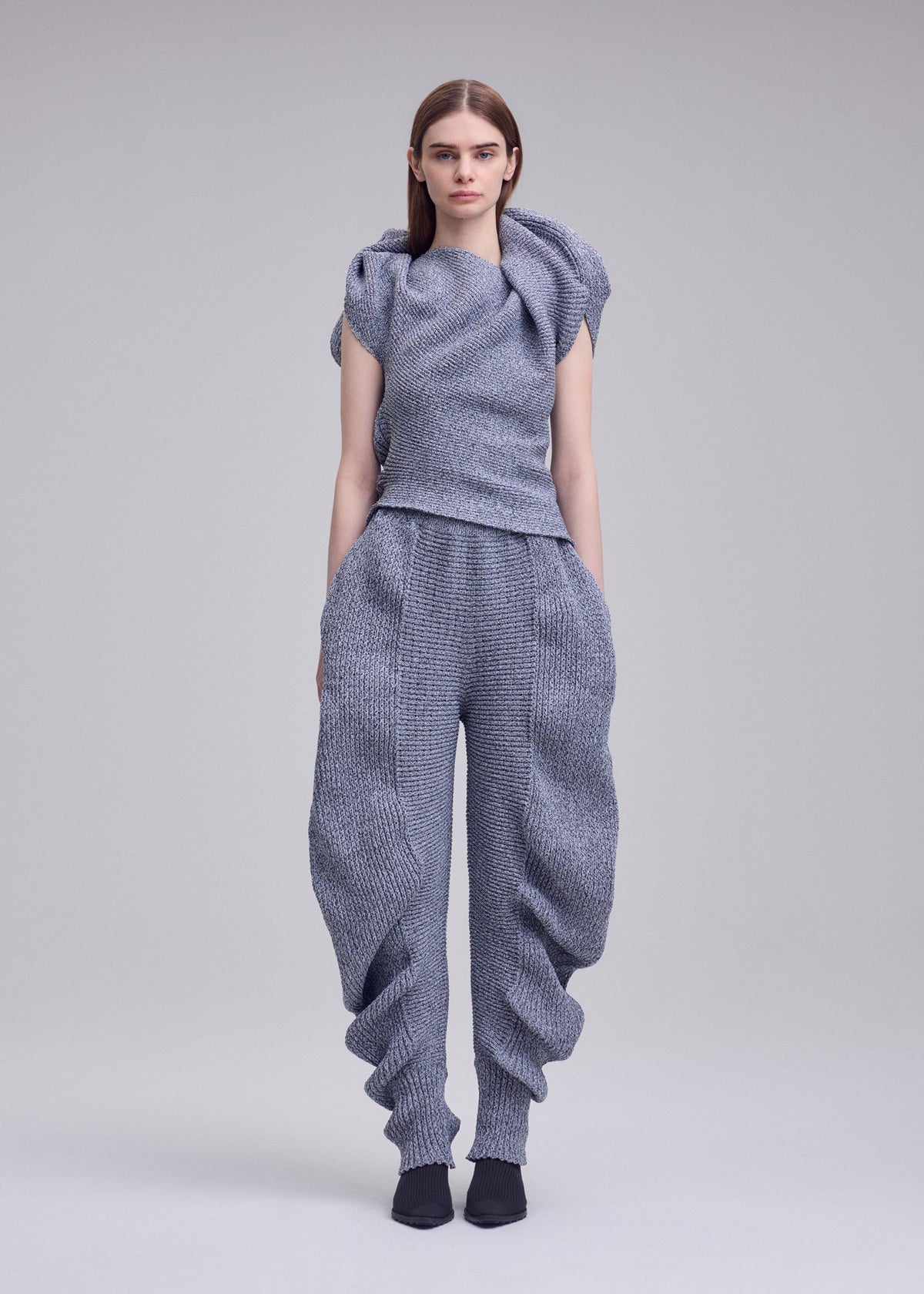 A person with straight brown hair and a neutral expression faces forward, wearing the FOUND STONE knit top by ISSEY MIYAKE with loose grey pants and black shoes, standing against a plain background. view 5 of 8