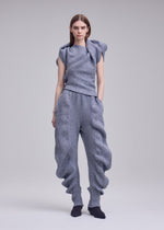 A woman stands against a plain background, wearing the ISSEY MIYAKE FOUND STONE set in light gray with sculptural, wavy details on the top and pants. She pairs it with black shoes and faces forward, arms relaxed by her sides.
