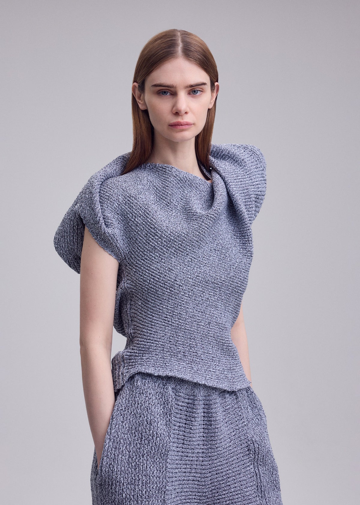 A woman with straight brown hair models ISSEY MIYAKE's FOUND STONE knit top, with a sculpted draped neckline and matching pants, posed against a plain light gray background. view 7 of 8
