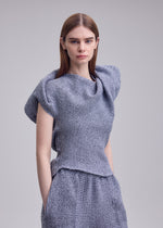 A woman with straight brown hair models ISSEY MIYAKE's FOUND STONE knit top, with a sculpted draped neckline and matching pants, posed against a plain light gray background.