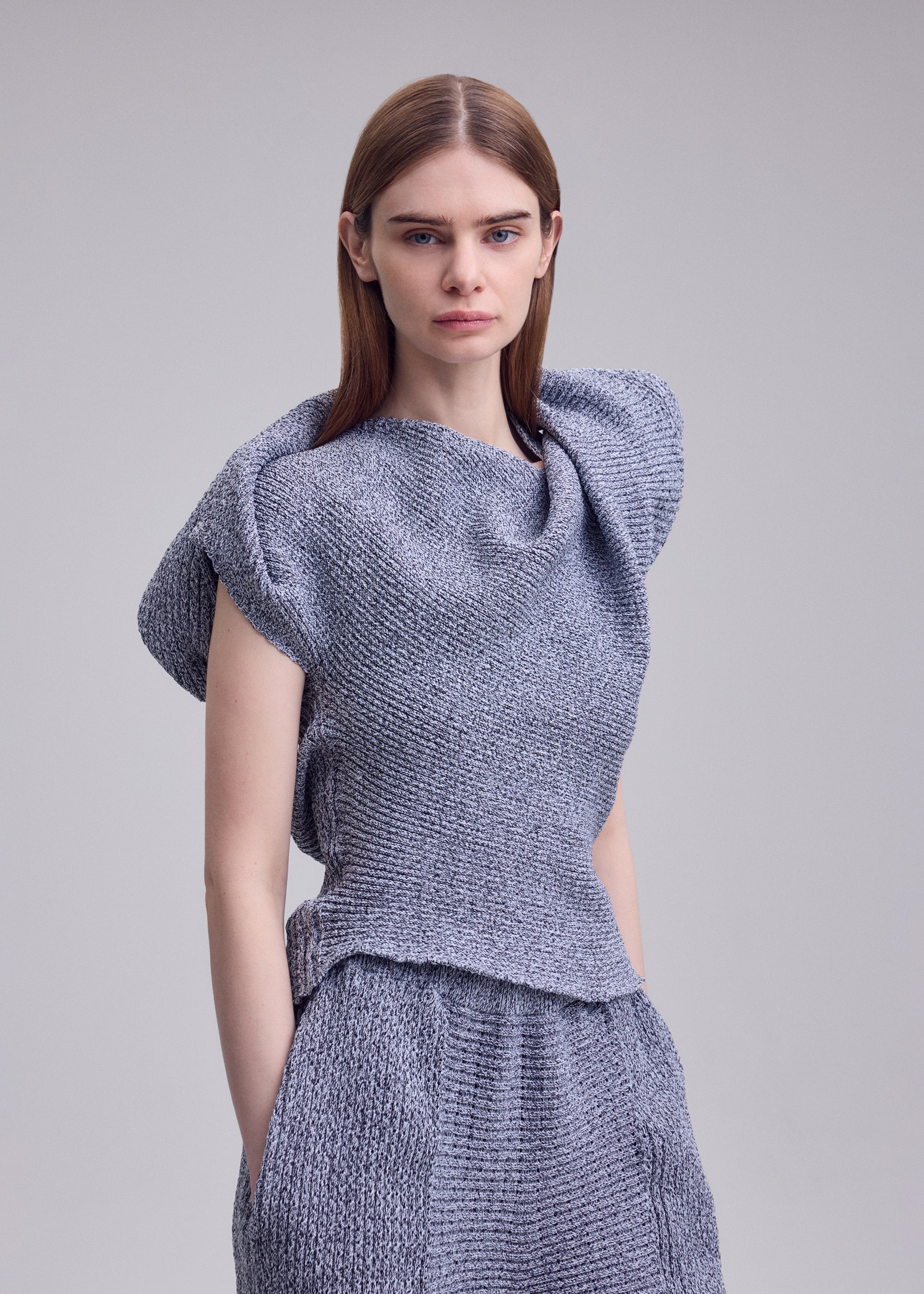 Zoomed view A woman with straight brown hair models ISSEY MIYAKE's FOUND STONE knit top, with a sculpted draped neckline and matching pants, posed against a plain light gray background.