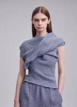 A person with straight, long brown hair wears the ISSEY MIYAKE FOUND STONE set—an organic, textured, asymmetrical light gray mesh knit top and matching pants—standing against a plain gray backdrop.