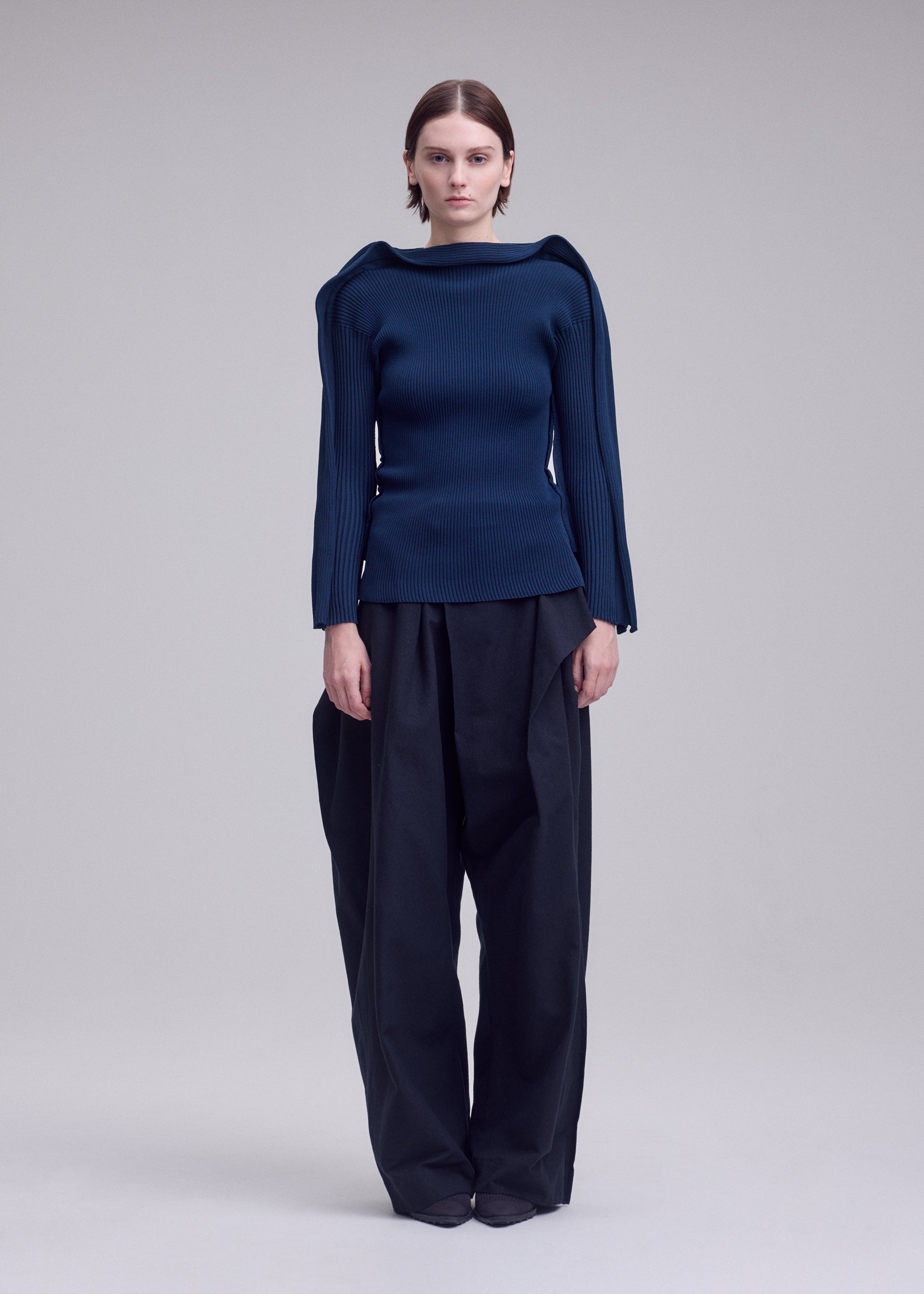 Zoomed view A person stands facing forward, wearing the ISSEY MIYAKE PRANCE navy blue long-sleeved knit top with structured shoulders and wide-legged black pants against a plain light gray background. The PRANCE sweater is crafted from recycled polyester yarn, combining sustainability with style.