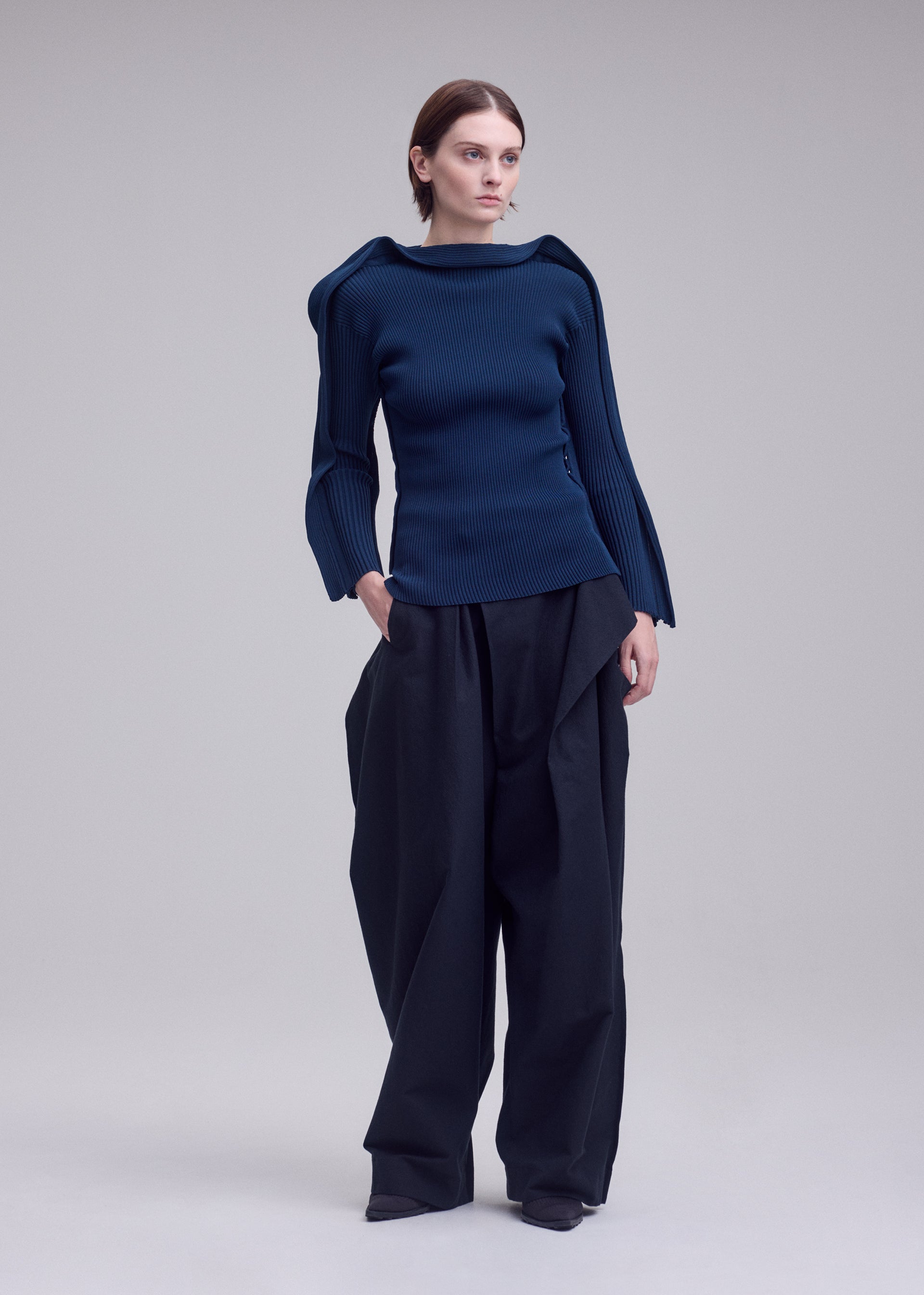 Zoomed view A person stands against a plain background, wearing ISSEY MIYAKE’s PRANCE—a fitted, long-sleeved blue knit top with sculpted shoulders—paired with wide-leg black pants. The ensemble exudes a modern, fashion-forward aesthetic, seamlessly blending sleek design and contemporary style.
