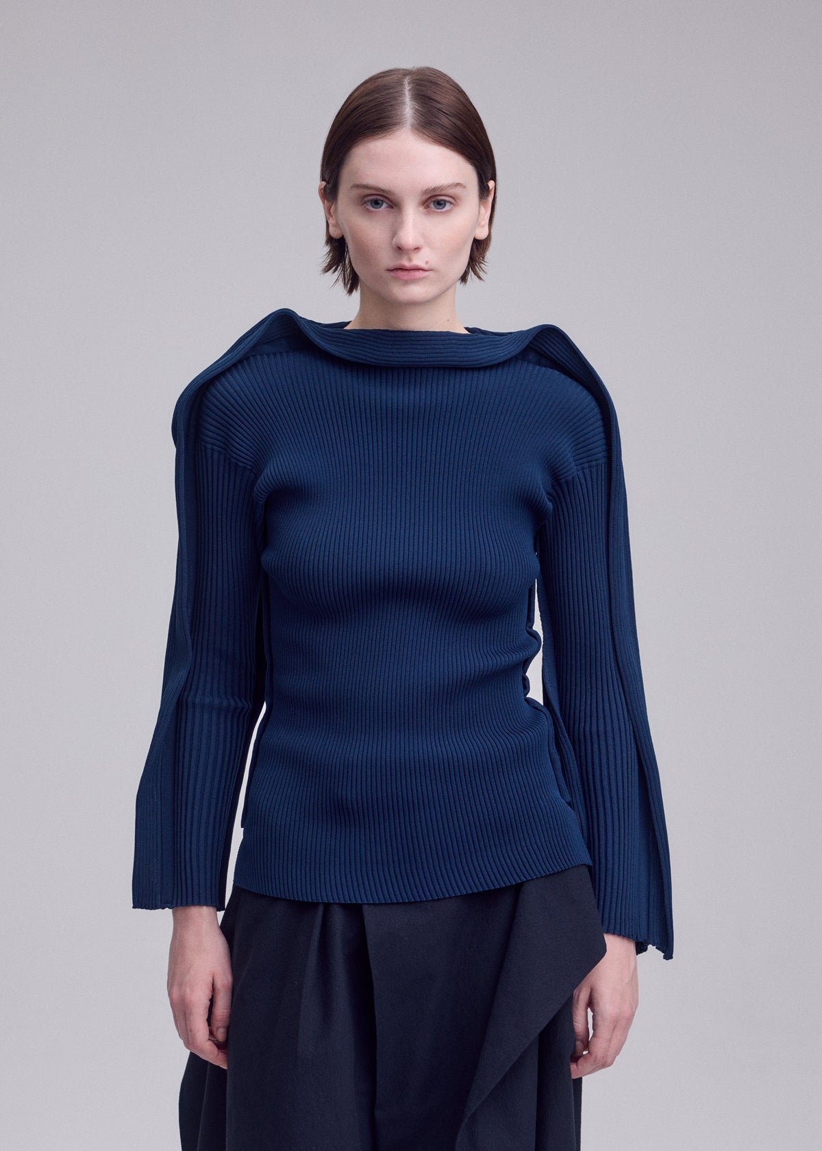 A person stands against a plain light background, looking directly at the camera while wearing PRANCE by ISSEY MIYAKE—a structured, dark blue long-sleeved knit top featuring unique shoulder details and a subtle quadrilateral outer frame pattern, crafted from recycled polyester yarn—paired with dark pants. view 7 of 7