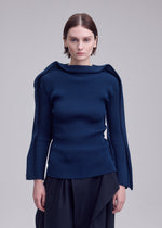 A person stands against a plain light background, looking directly at the camera while wearing PRANCE by ISSEY MIYAKE—a structured, dark blue long-sleeved knit top featuring unique shoulder details and a subtle quadrilateral outer frame pattern, crafted from recycled polyester yarn—paired with dark pants.