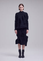 A woman stands facing forward against a plain light gray background, dressed in the ISSEY MIYAKE KONE KONE textured knit long-sleeved black top and matching skirt, paired with mid-calf black socks and black heels.