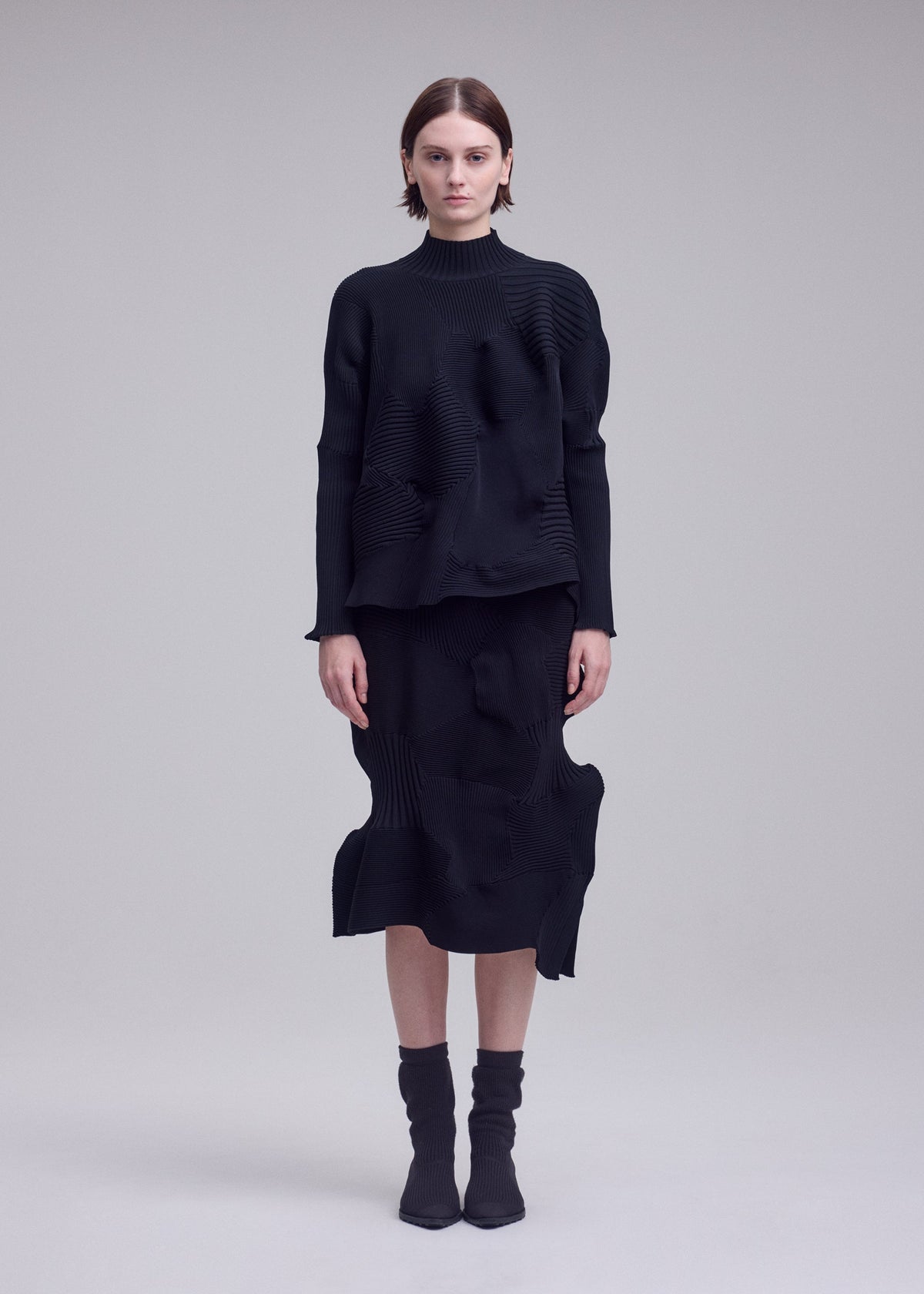 Zoomed view A woman stands facing forward against a plain light gray background, dressed in the ISSEY MIYAKE KONE KONE textured knit long-sleeved black top and matching skirt, paired with mid-calf black socks and black heels.