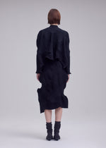 A person with short brown hair stands facing away, dressed in the ISSEY MIYAKE KONE KONE black textured knit top and a flowing midi skirt. They are also wearing black socks and pointed shoes, set against a plain, light-colored background.