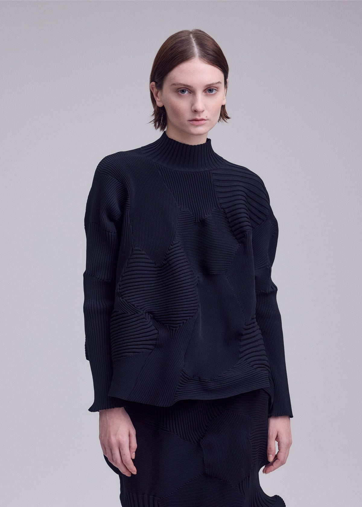 A person with short brown hair wears the ISSEY MIYAKE KONE KONE textured knit long-sleeve top and matching skirt, standing against a plain light gray background. view 6 of 6