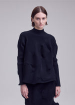 A person with short brown hair wears the ISSEY MIYAKE KONE KONE textured knit long-sleeve top and matching skirt, standing against a plain light gray background.