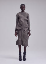 A person stands against a plain background, wearing the ISSEY MIYAKE KONE KONE textured knit earth-toned long-sleeve top and matching ribbed midi skirt, paired with black ankle boots.