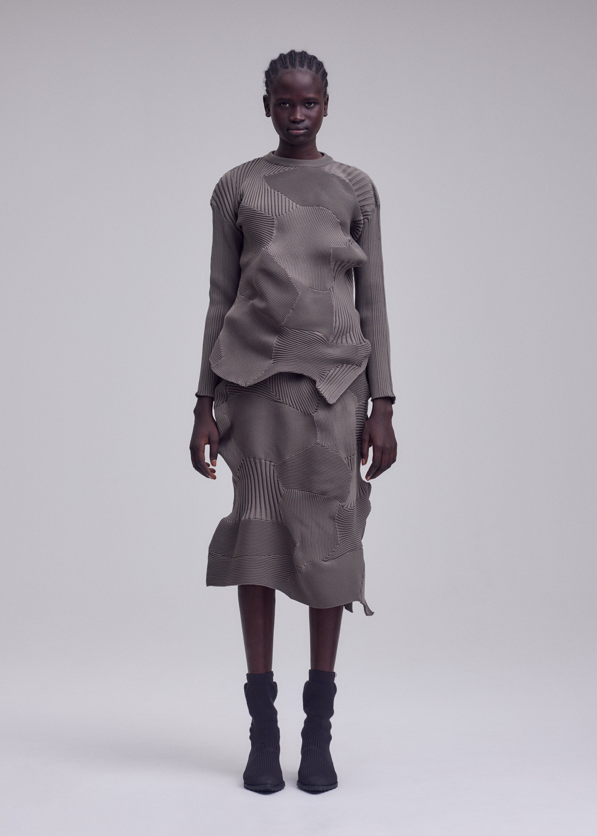 Zoomed view A person stands against a plain background, wearing the ISSEY MIYAKE KONE KONE textured knit earth-toned long-sleeve top and matching ribbed midi skirt, paired with black ankle boots.