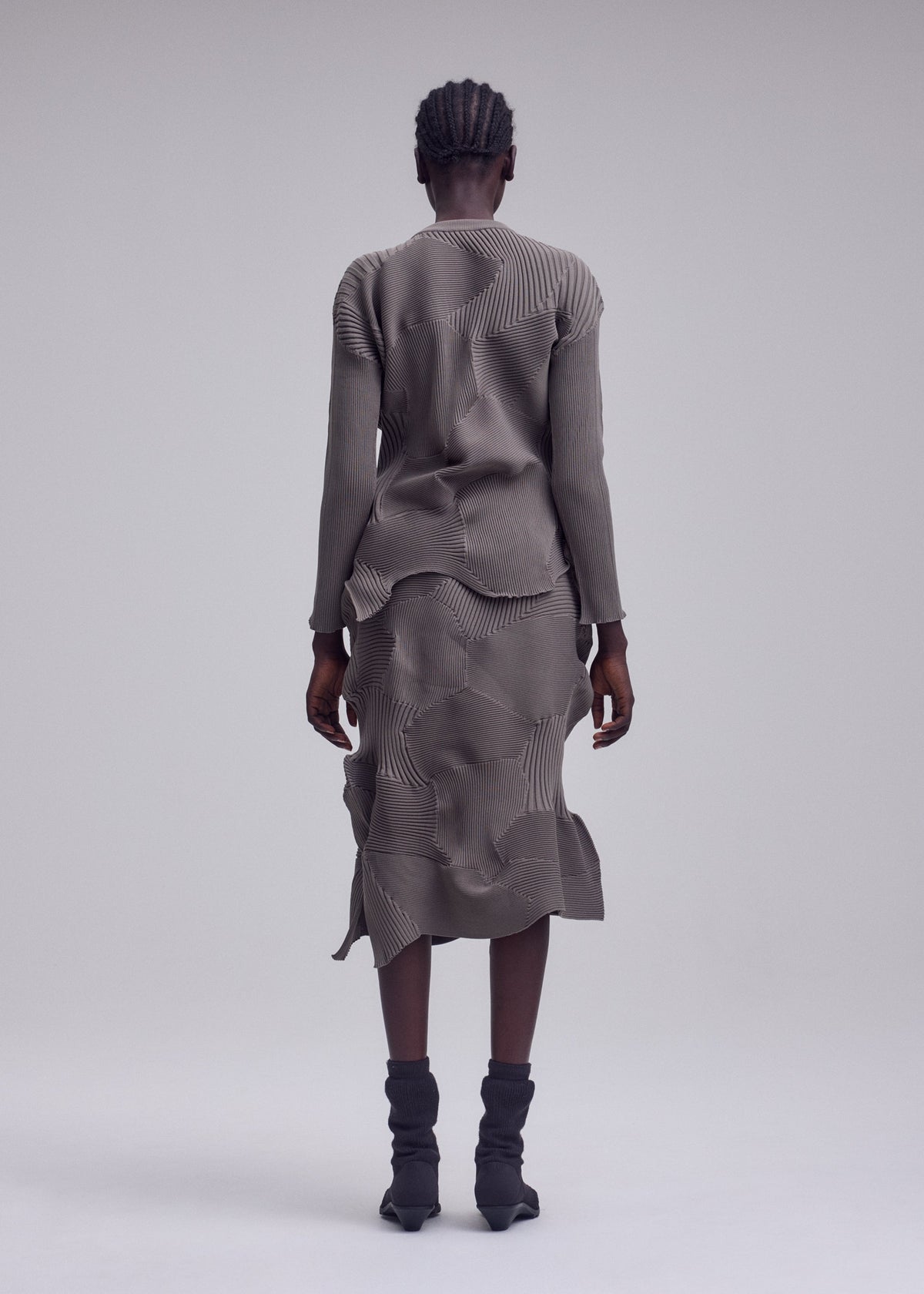 A person with short braided hair stands facing away, wearing the KONE KONE textured knit gray long-sleeved top and matching skirt by ISSEY MIYAKE, paired with black mid-calf boots against a plain light gray background. view 4 of 5
