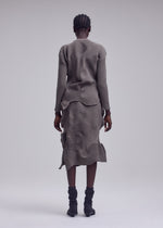 A person with short braided hair stands facing away, wearing the KONE KONE textured knit gray long-sleeved top and matching skirt by ISSEY MIYAKE, paired with black mid-calf boots against a plain light gray background.