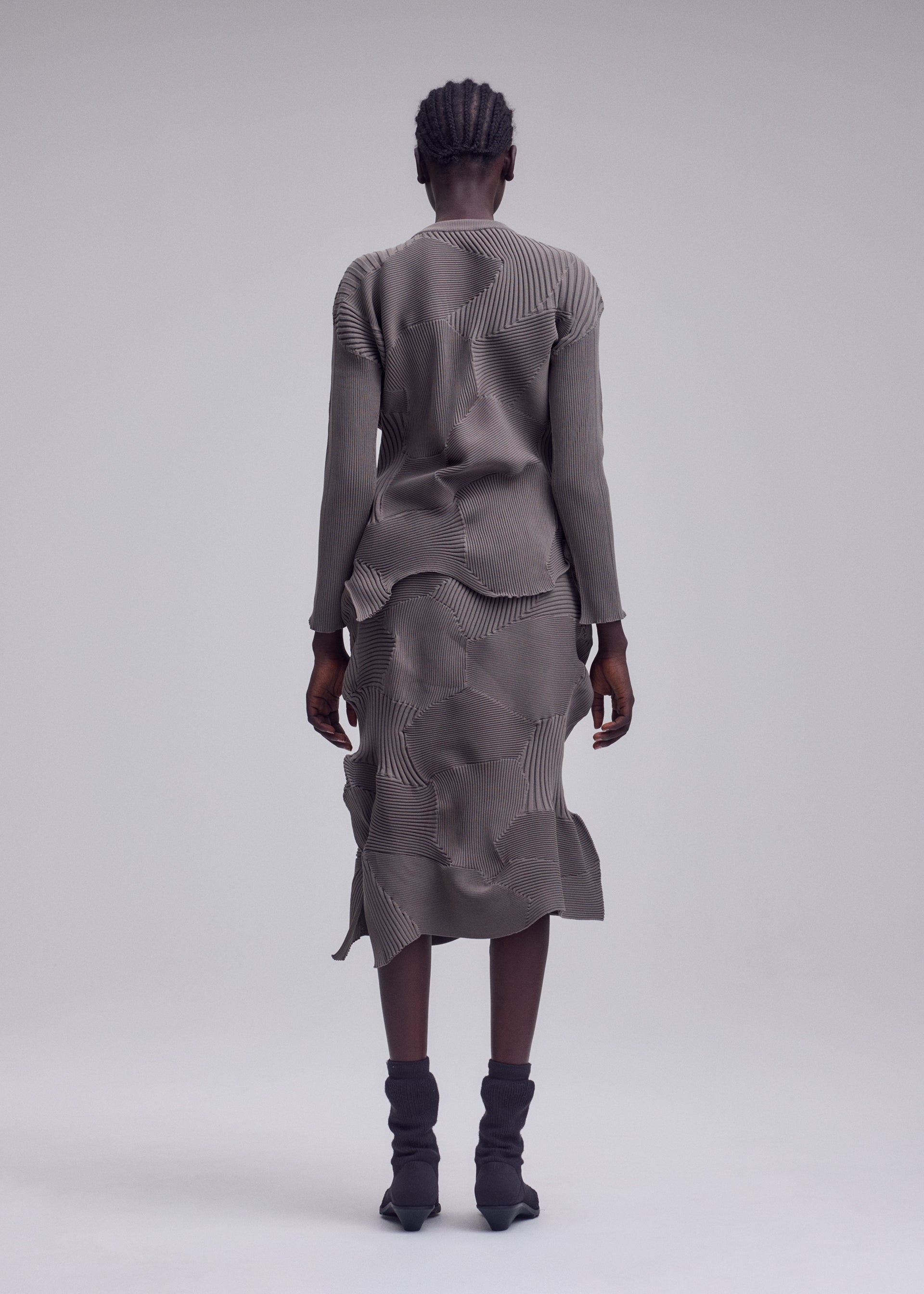 Zoomed view A person with short braided hair stands facing away, wearing the KONE KONE textured knit gray long-sleeved top and matching skirt by ISSEY MIYAKE, paired with black mid-calf boots against a plain light gray background.