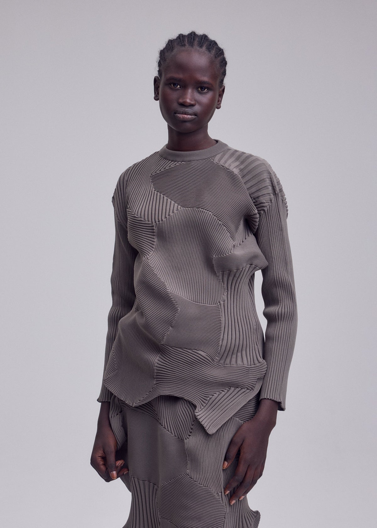 A person with short braids wears the ISSEY MIYAKE KONE KONE textured knit patterned taupe long-sleeved top and matching skirt, standing against a plain gray background. view 5 of 5