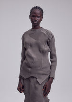 A person with short braids wears the ISSEY MIYAKE KONE KONE textured knit patterned taupe long-sleeved top and matching skirt, standing against a plain gray background.