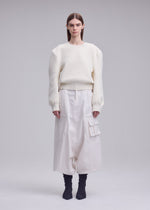 A person stands facing forward in an ISSEY MIYAKE HANDSOME KNIT white oversized sweater with 3D shoulder detail and wide white cargo pants, straight brown hair, and black ankle boots against a light gray background.