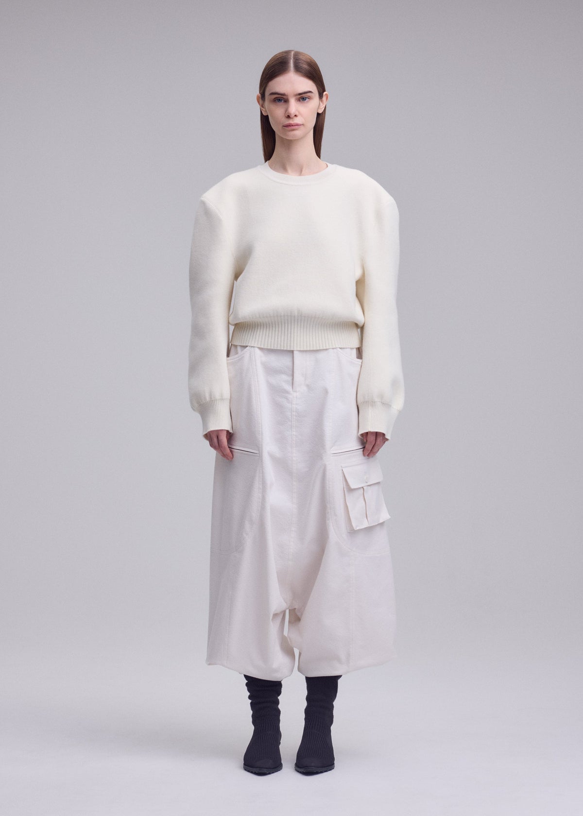 Zoomed view A person stands facing forward in an ISSEY MIYAKE HANDSOME KNIT white oversized sweater with 3D shoulder detail and wide white cargo pants, straight brown hair, and black ankle boots against a light gray background.