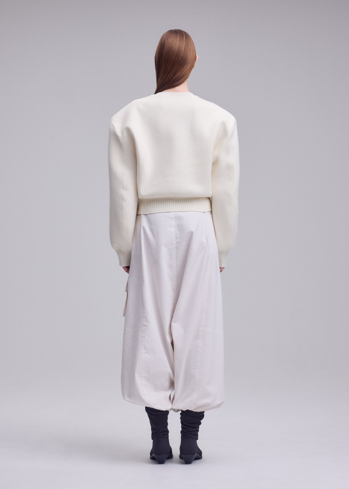 A person with long, straight hair faces away, wearing the ISSEY MIYAKE HANDSOME KNIT, loose white pants, and black ankle boots against a plain gray background. view 5 of 6