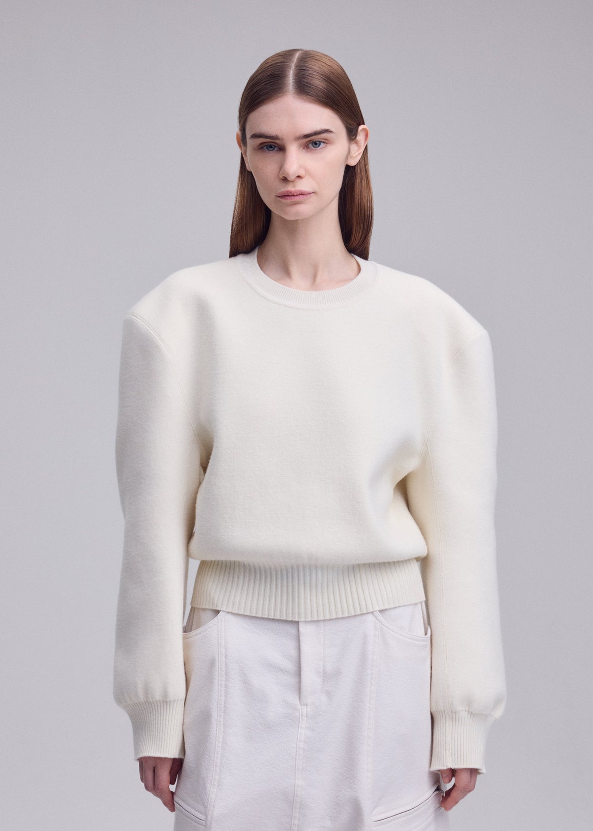 Zoomed view A woman with straight brown hair wears an oversized ISSEY MIYAKE HANDSOME KNIT white sweater and white skirt with three-dimensional shoulders, standing against a plain light gray background.