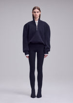 A person stands against a plain background in the ISSEY MIYAKE HANDSOME KNIT with ribbed leggings and black shoes. Their straight hair is tucked behind their ears, and they have a neutral expression.