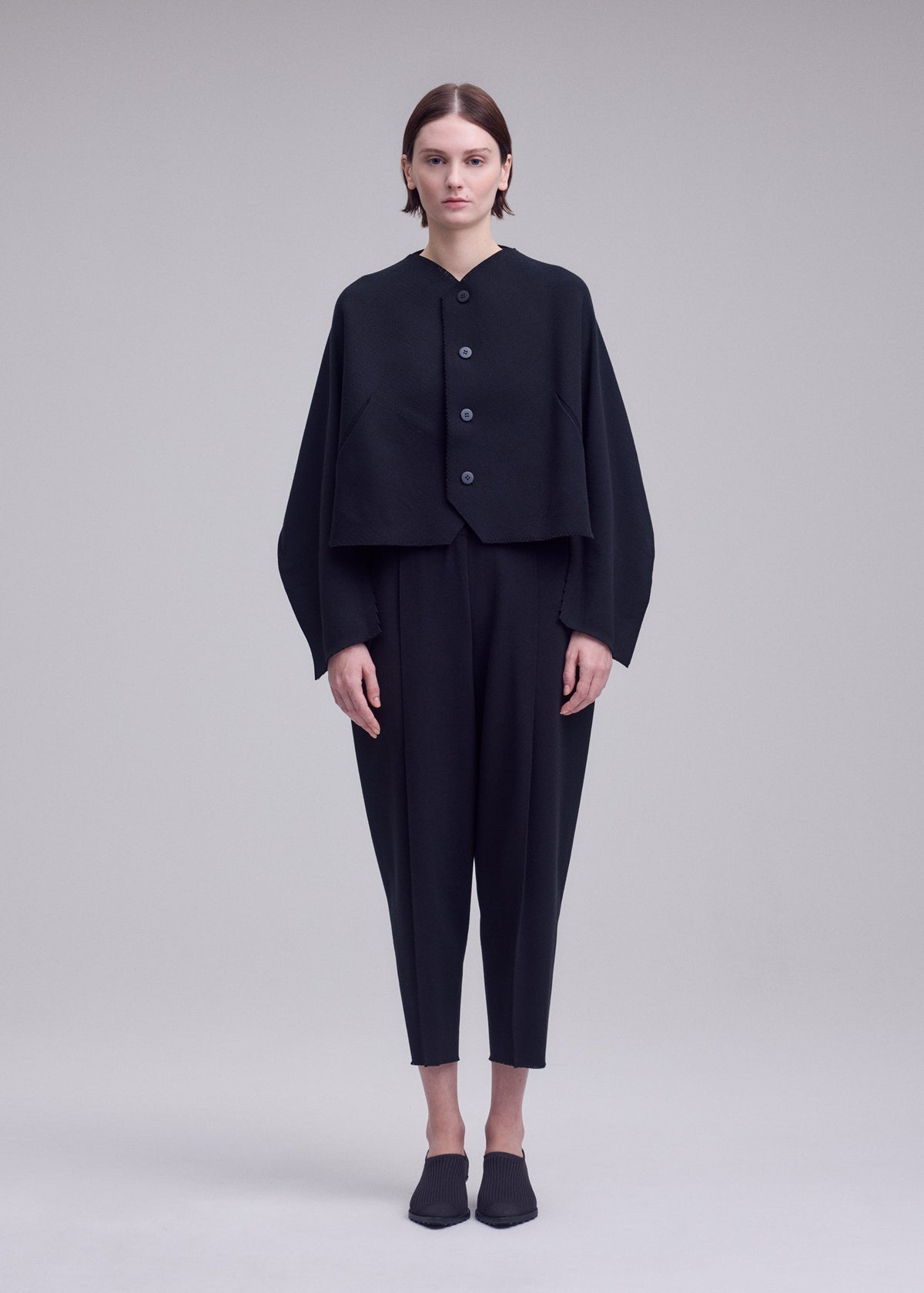 A person stands facing forward against a plain light grey background, wearing ISSEY MIYAKE’s CAMPAGNE ensemble: a loose black buttoned jacket crafted from innovative A-POC material, paired with matching black cropped pants and black shoes. view 3 of 5