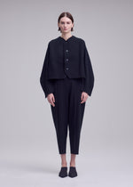 A person stands facing forward against a plain light grey background, wearing ISSEY MIYAKE’s CAMPAGNE ensemble: a loose black buttoned jacket crafted from innovative A-POC material, paired with matching black cropped pants and black shoes.
