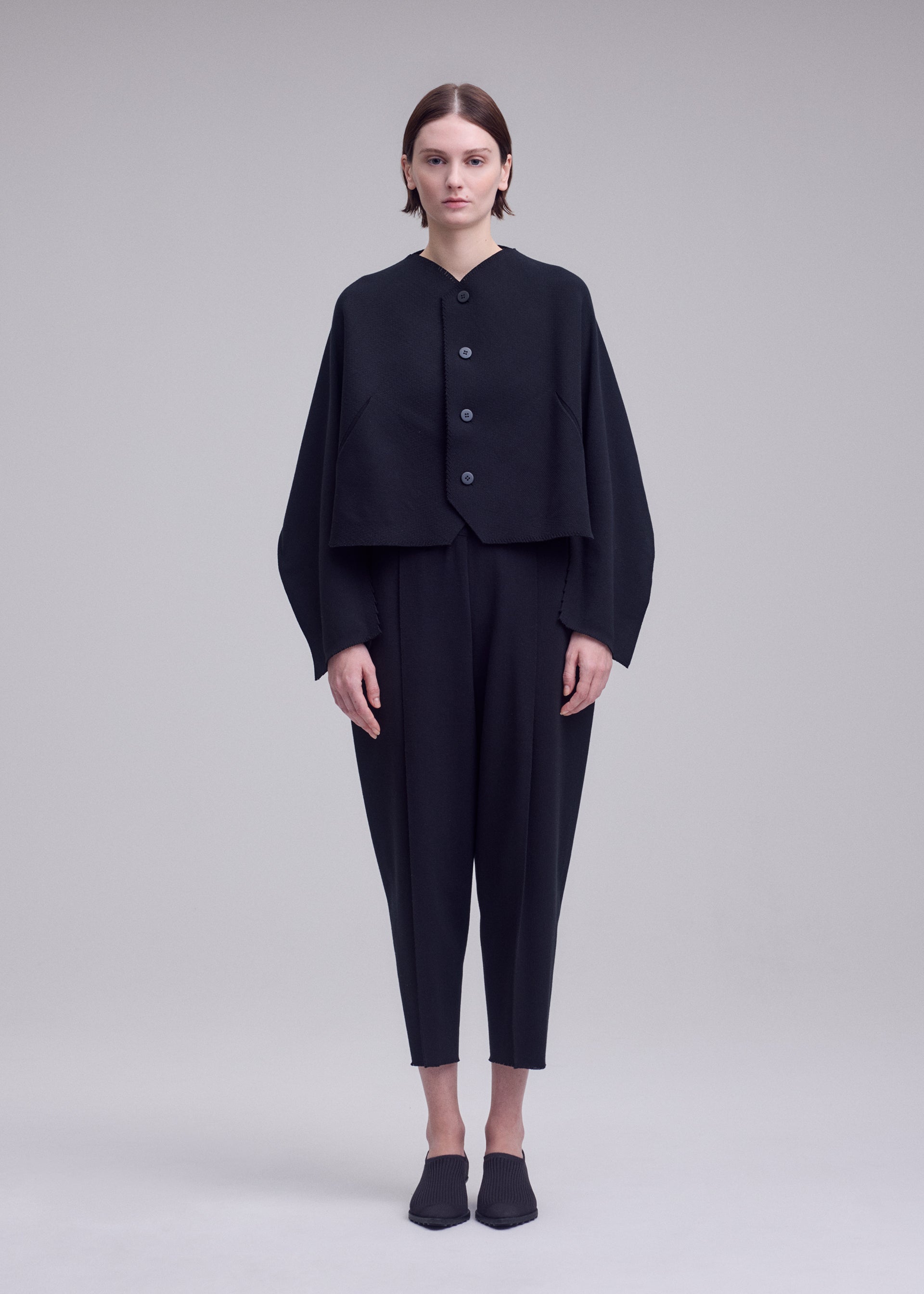Zoomed view A person stands facing forward against a plain light grey background, wearing ISSEY MIYAKE’s CAMPAGNE ensemble: a loose black buttoned jacket crafted from innovative A-POC material, paired with matching black cropped pants and black shoes.