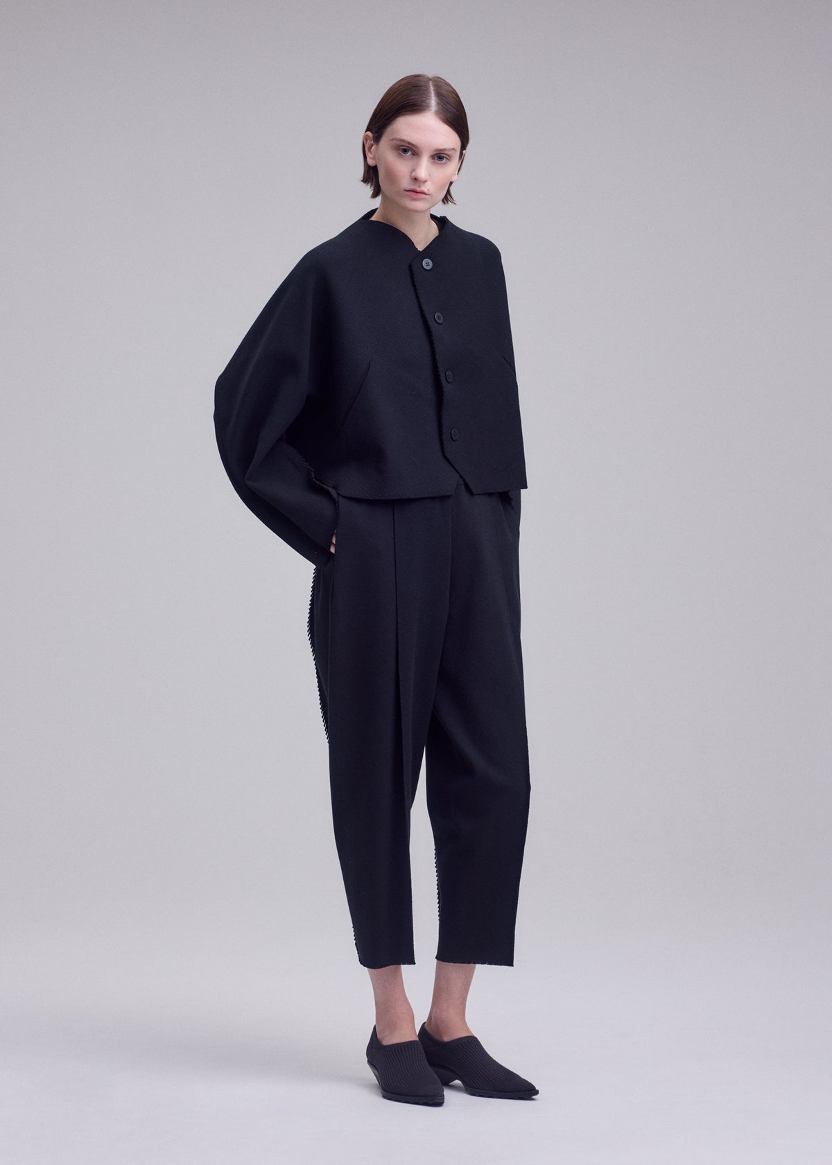 A person stands against a plain background wearing the ISSEY MIYAKE CAMPAGNE jacket, a loose black buttoned piece crafted from signature A-POC material, paired with matching black pants and black slip-on shoes, hands in pockets and sporting a neutral expression. view 4 of 5