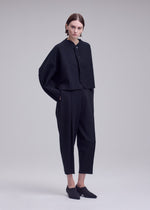 A person stands against a plain background wearing the ISSEY MIYAKE CAMPAGNE jacket, a loose black buttoned piece crafted from signature A-POC material, paired with matching black pants and black slip-on shoes, hands in pockets and sporting a neutral expression.
