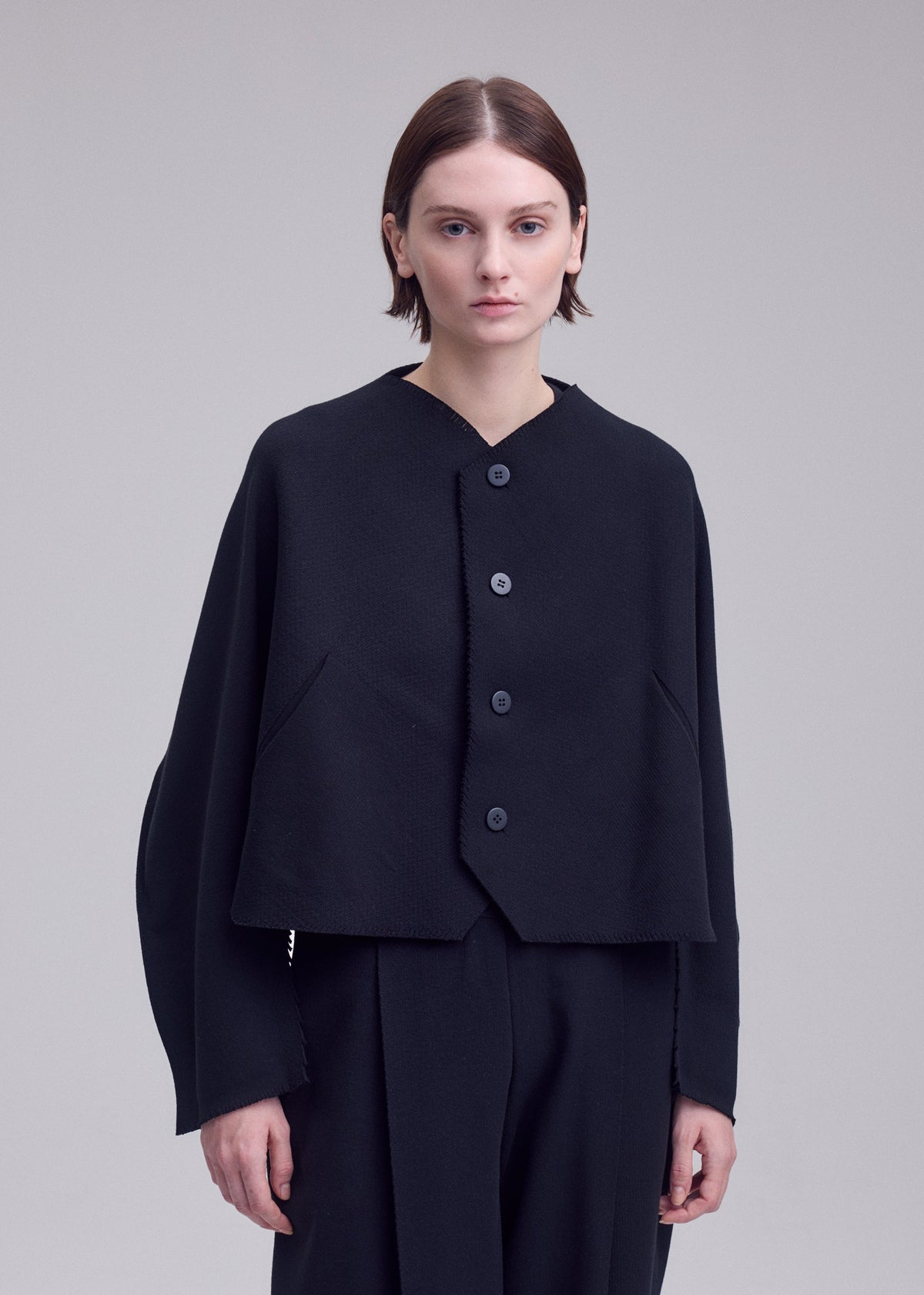 A person with short brown hair wears the ISSEY MIYAKE CAMPAGNE black buttoned jacket, crafted from innovative A-POC material, along with matching black pants, standing against a plain light gray background. view 5 of 5